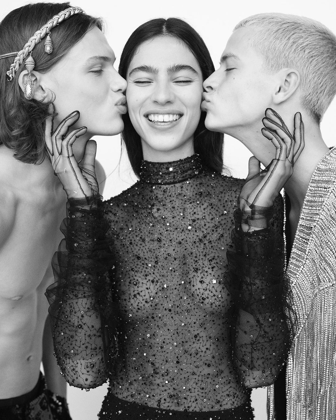 Black and white fashion photo of a smiling woman in a beaded top getting kissed on the cheeks by two men in a bright indoor space.