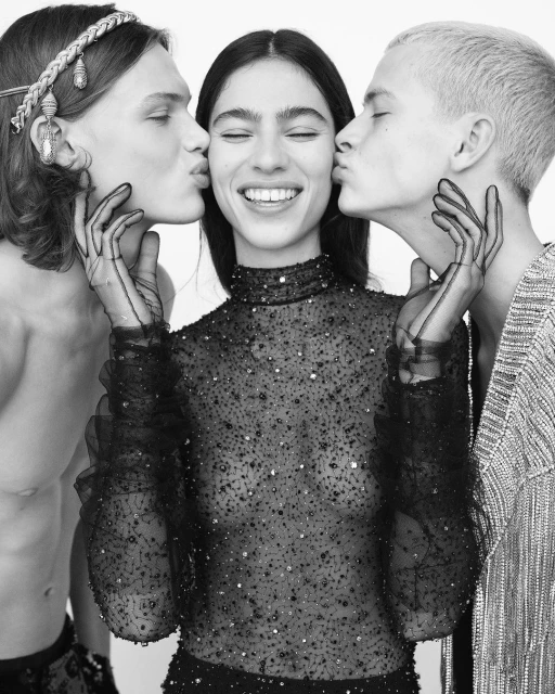 Black and white fashion photo of a smiling woman in a beaded top getting kissed on the cheeks by two men in a bright indoor space.