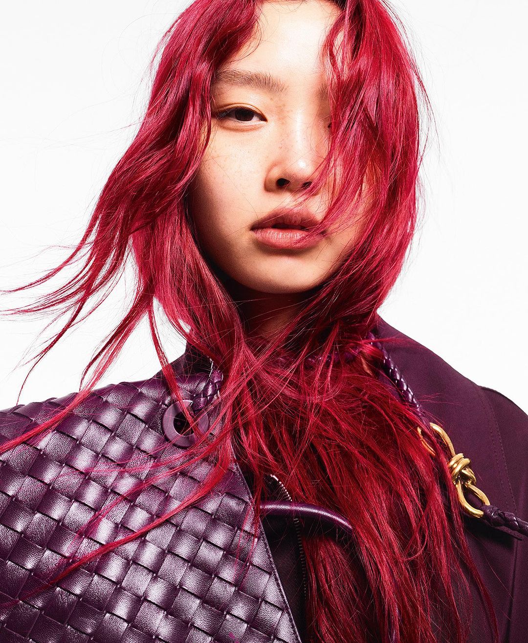 Close-up of a person with vibrant red hair, wearing a dark jacket and carrying a woven leather bag, posing in a brightly lit studio.