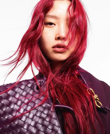 Close-up of a person with vibrant red hair, wearing a dark jacket and carrying a woven leather bag, posing in a brightly lit studio.