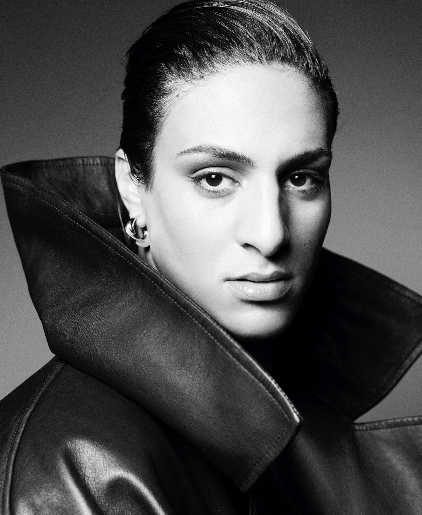Close-up monochrome portrait of a person with slicked-back dark hair wearing a high-collared leather coat by Karim Sadli.
