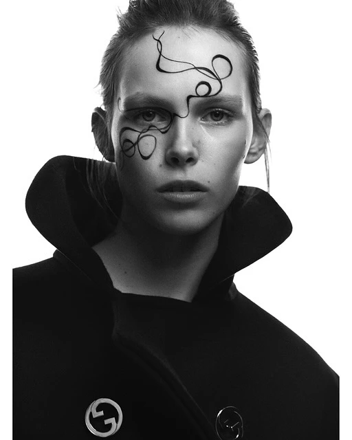 Close-up portrait of a woman with stylized makeup art on her face and a dark coat against a bright background in a black and white photo.
