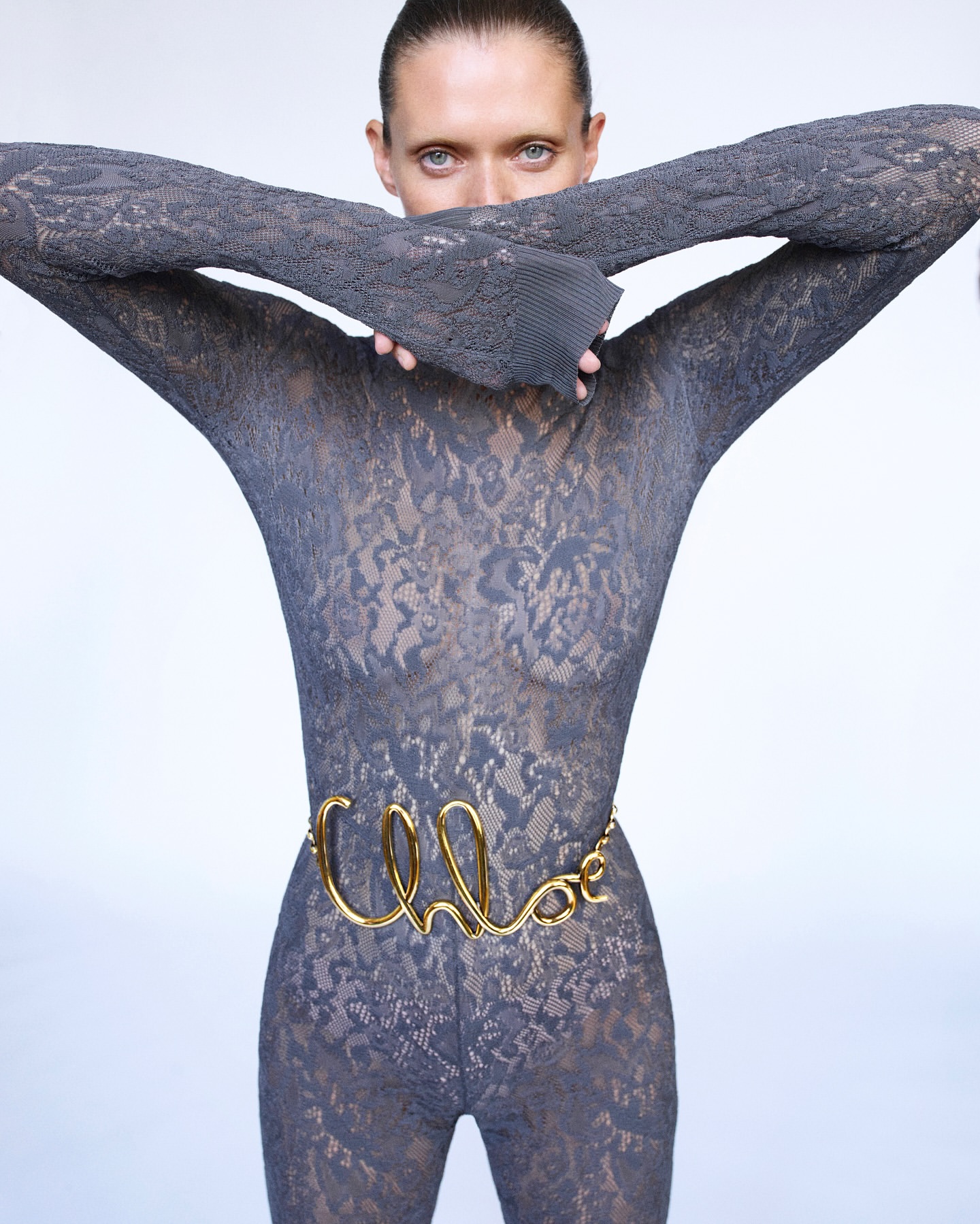 A slim, fair-skinned person poses in a lace catsuit with a golden belt and hands raised, set against a stark white studio backdrop.