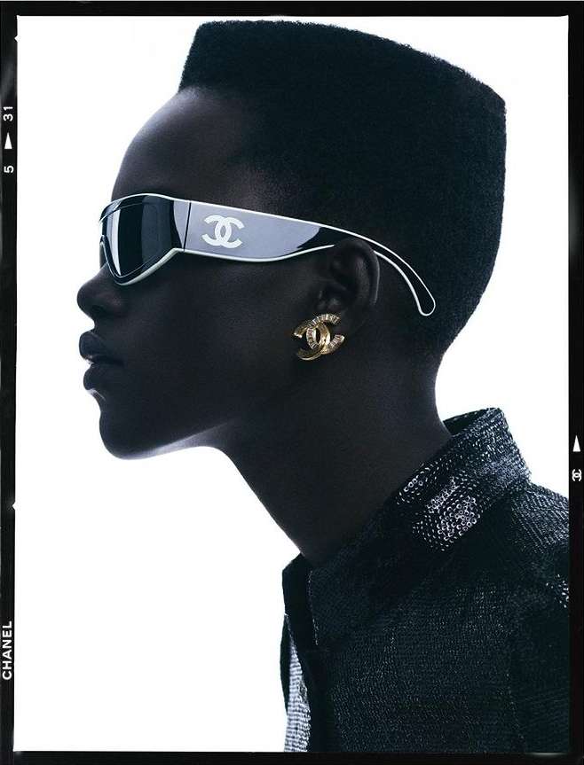 Profile shot of a Black adult female wearing designer sunglasses and jewelry, indoors against a white backdrop, framed by dark edges.