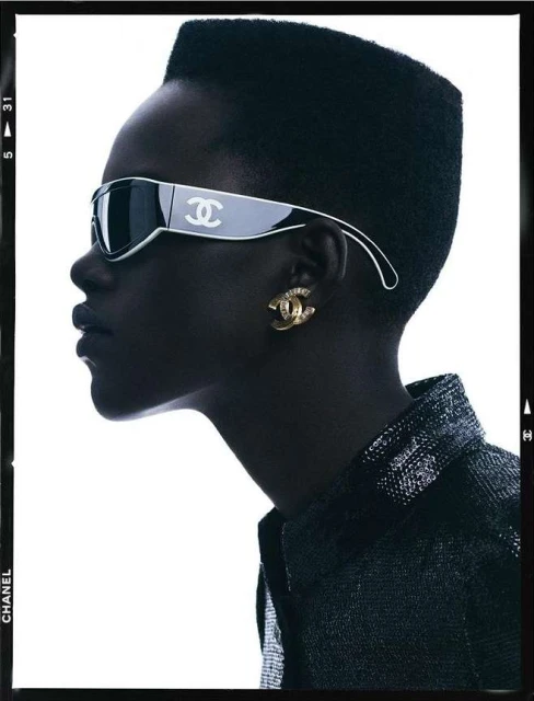 Profile shot of a Black adult female wearing designer sunglasses and jewelry, indoors against a white backdrop, framed by dark edges.