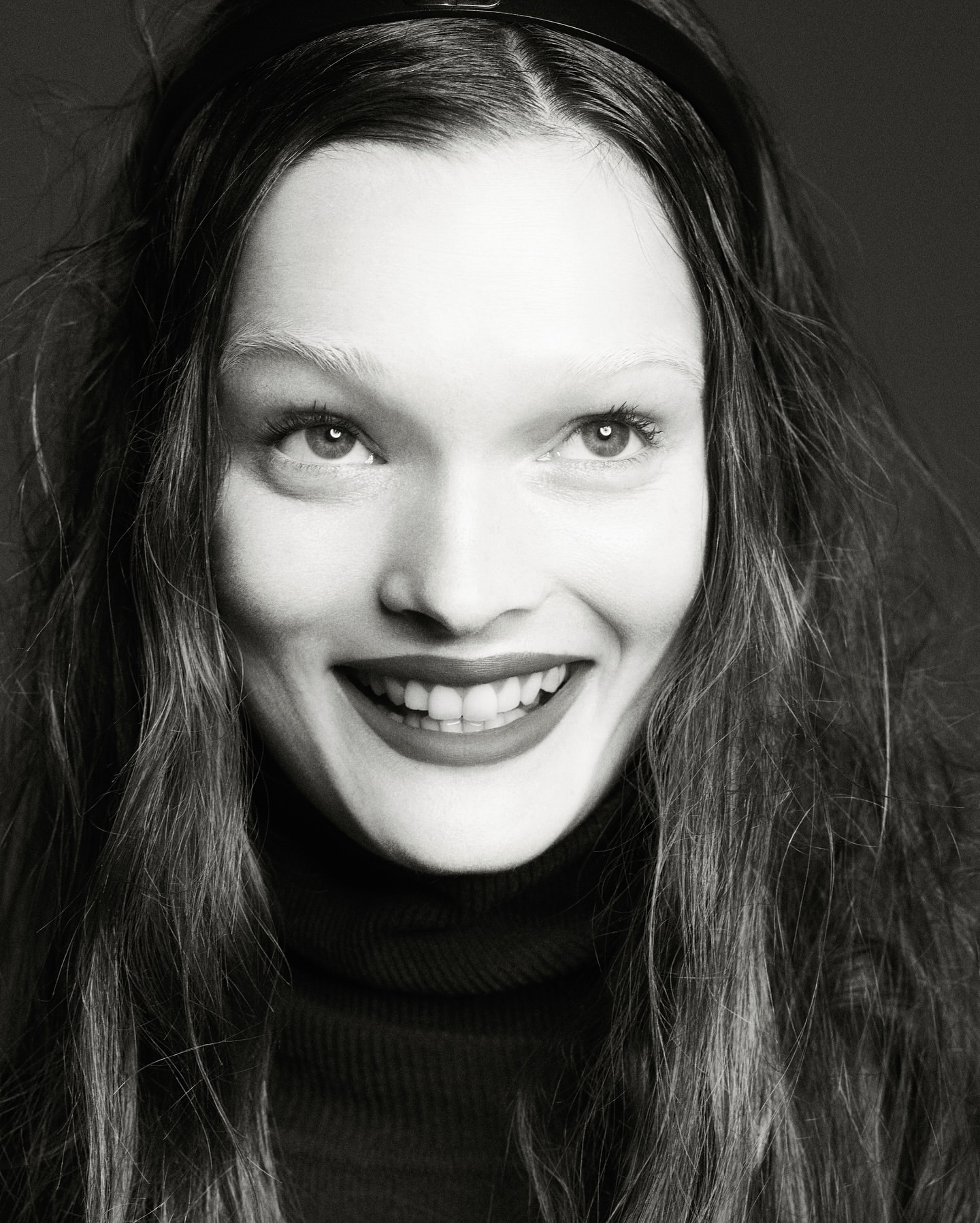Close-up portrait in black and white of a smiling adult woman with long hair and a headband, wearing a dark turtleneck sweater.