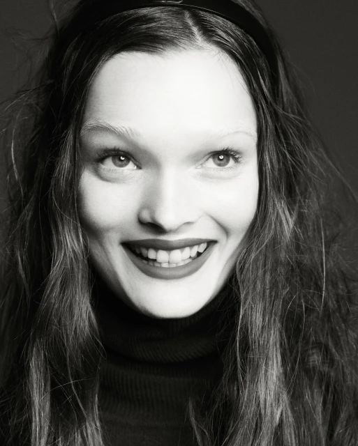 Close-up portrait in black and white of a smiling adult woman with long hair and a headband, wearing a dark turtleneck sweater.