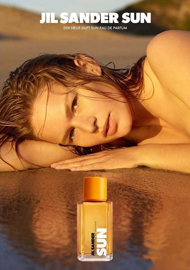 Sun-kissed skin radiates as a young woman reclines on a sandy beach beside a bottle of Jil Sander Sun perfume in a beauty editorial.