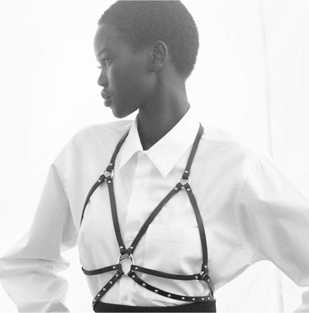 Portrait of a woman in a white shirt wearing a black harness, shot in black and white with diffused lighting against a white background.