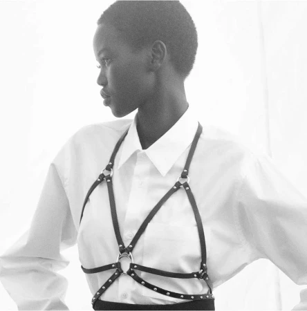 Portrait of a woman in a white shirt wearing a black harness, shot in black and white with diffused lighting against a white background.