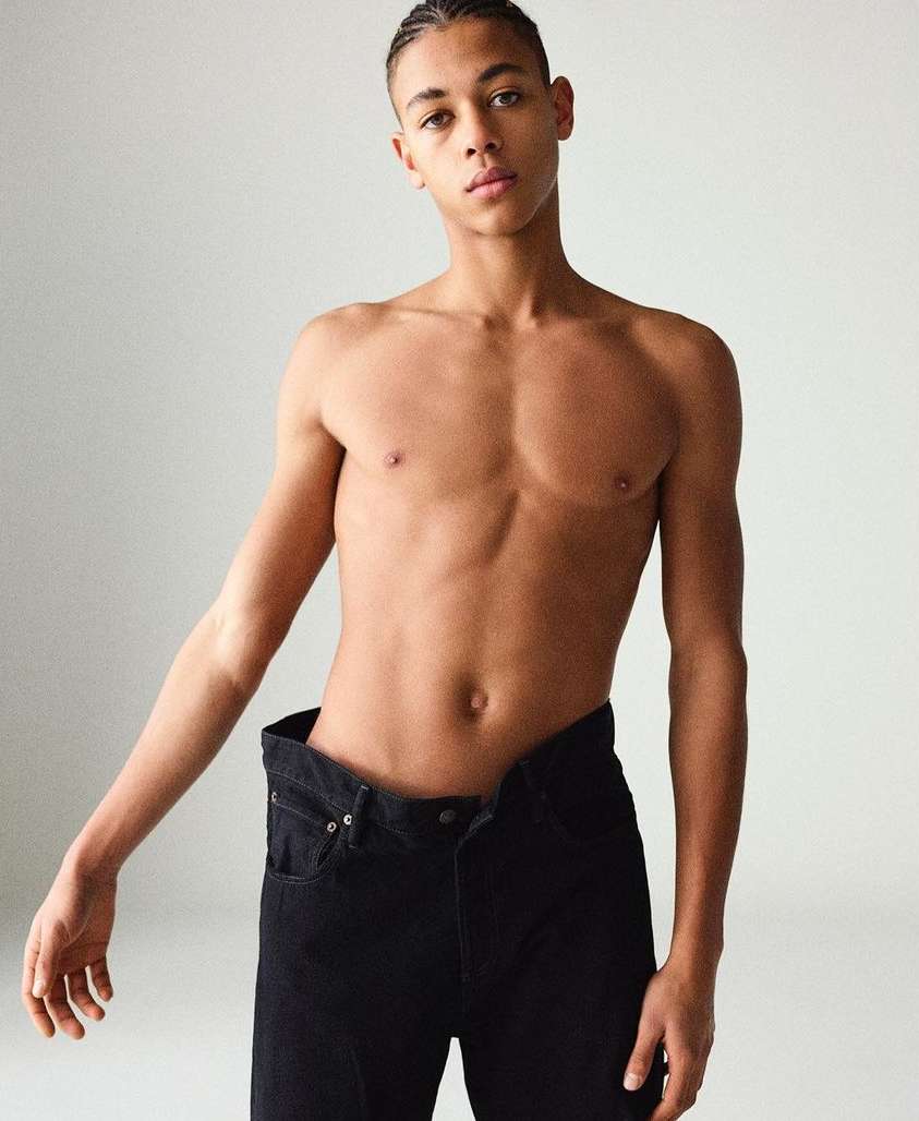 Portrait of shirtless male fashion model with braids, wearing black jeans against a plain off-white backdrop in a photo studio.