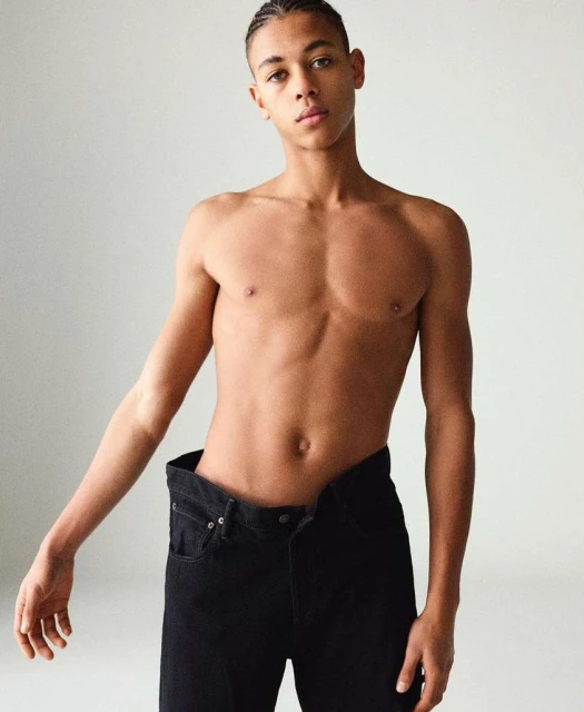 Portrait of shirtless male fashion model with braids, wearing black jeans against a plain off-white backdrop in a photo studio.