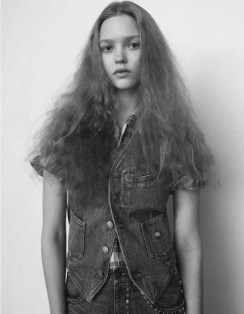 Portrait of a young woman with long wavy hair wearing a denim vest and plaid shirt against a plain wall.