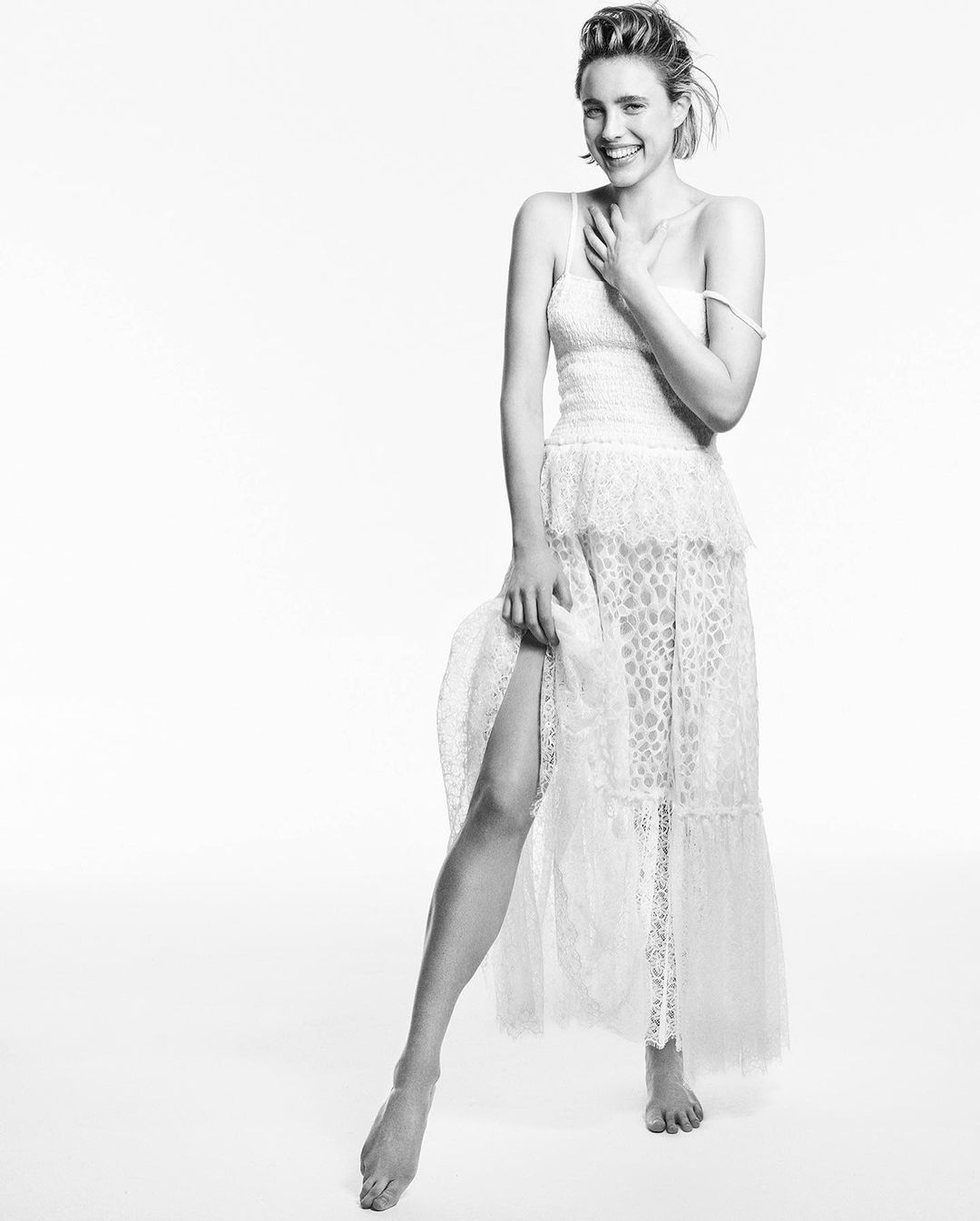 A smiling young adult female posing in a white lace dress against a bright white backdrop in a studio fashion shot.