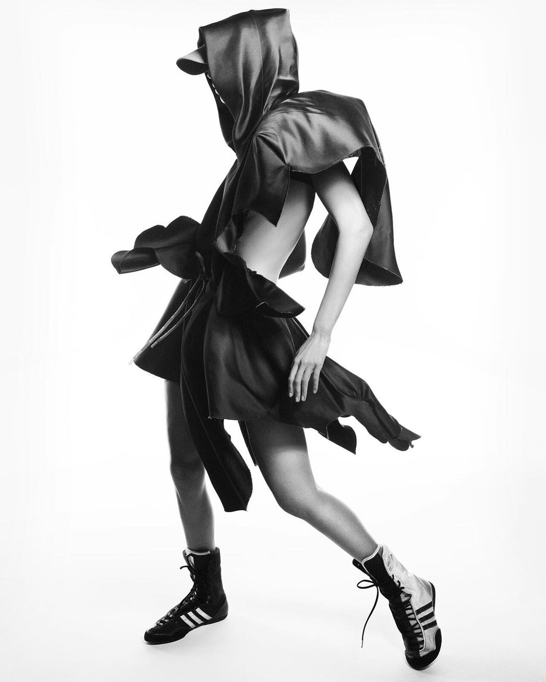 Monochrome fashion shot of a woman in an oversized hooded garment with boxing shoes against a white backdrop in a studio setting.