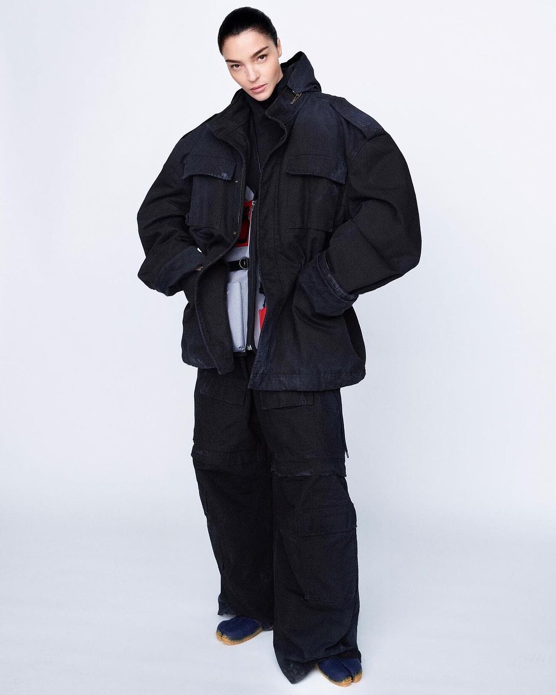 Full-length shot of a young adult female fashion model in oversized dark blue jacket, pants, and tabi shoes against a white backdrop.