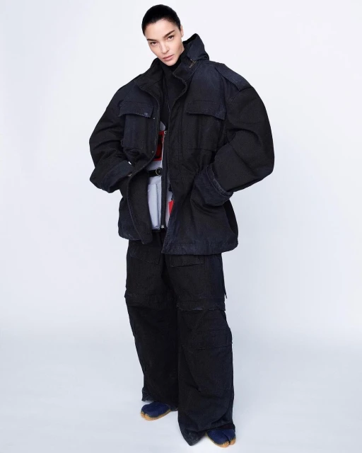 Full-length shot of a young adult female fashion model in oversized dark blue jacket, pants, and tabi shoes against a white backdrop.
