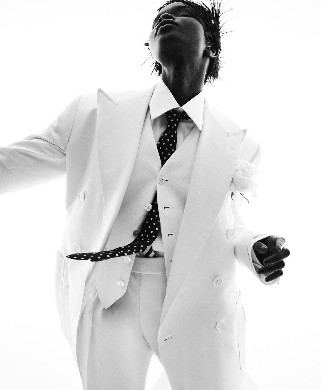 Black model in a white double-breasted suit and black polka dot tie strikes a pose against a bright white background in a fashion editorial.