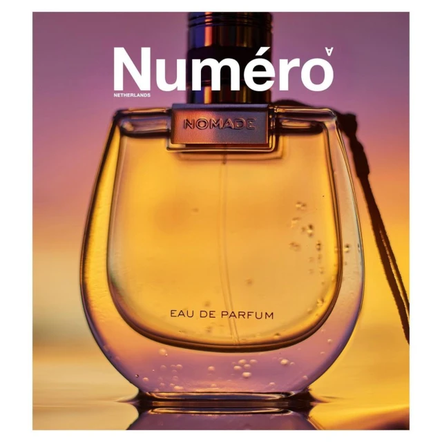 Glass perfume bottle of Nomade Eau de Parfum, with a gold liquid inside, on a reflective surface, and the brand logo Numero on top.