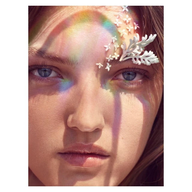 Close-up portrait of a young woman with light skin, blue eyes, rainbow light effect, and white flower accents in her hair. Beauty shot.