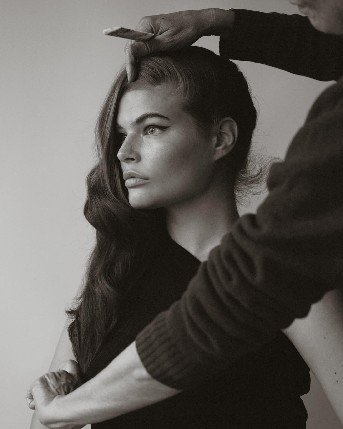 Monochrome portrait of a woman with long wavy hair having her hair styled by a hairstylist in an indoor setting.