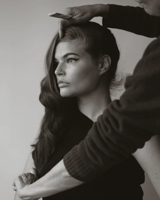 Monochrome portrait of a woman with long wavy hair having her hair styled by a hairstylist in an indoor setting.