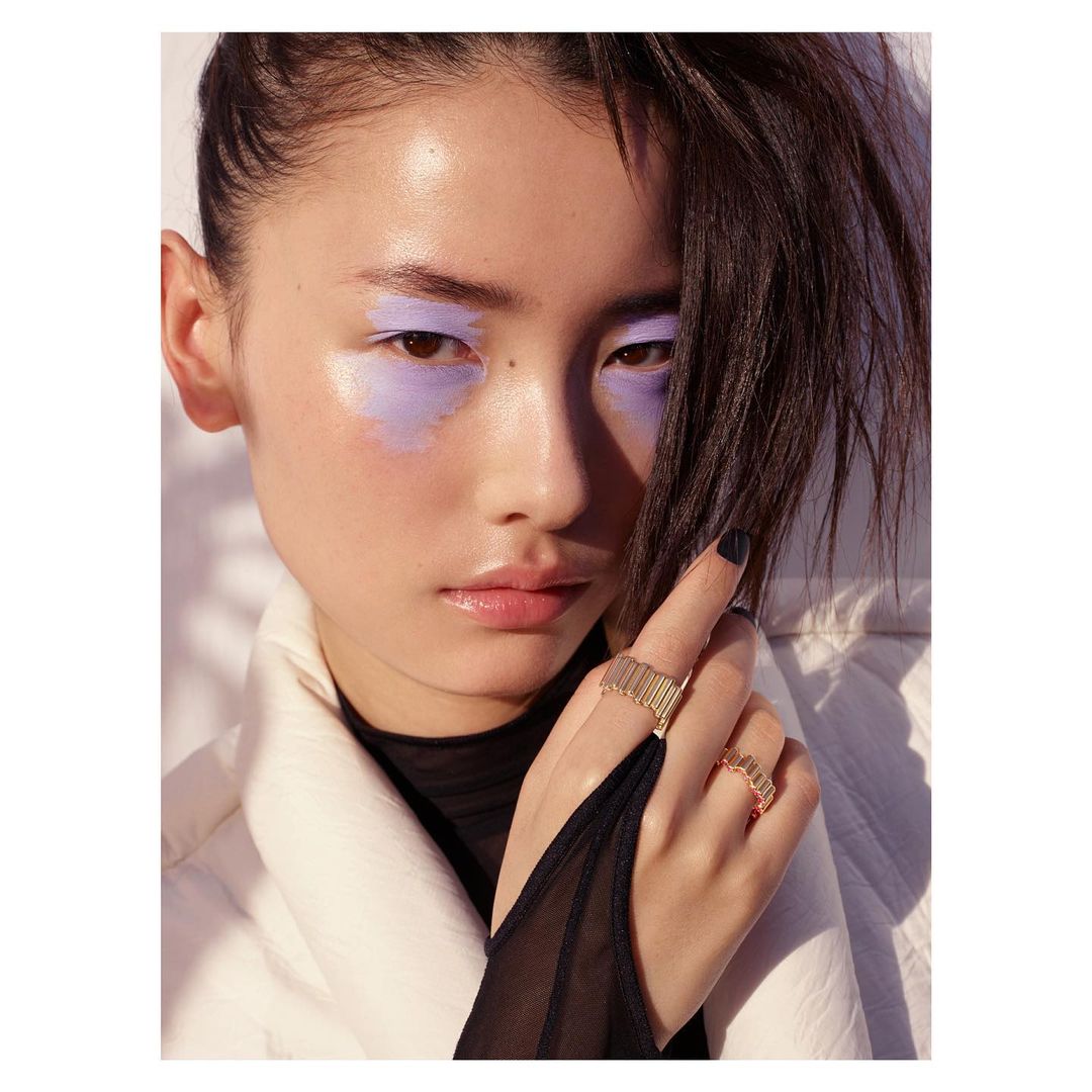 Close-up of a woman with light purple eyeshadow wearing gold and pinky-gold rings on her fingers in a fashion editorial shot.