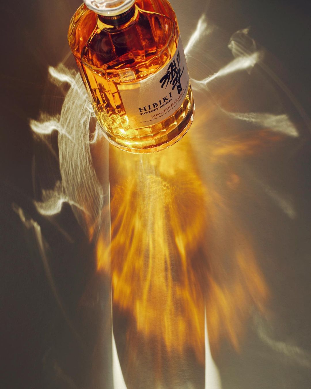 A glass bottle of Hibiki Suntory Whisky sits indoors, casting warm light and reflections on a smooth surface with neutral coloring.