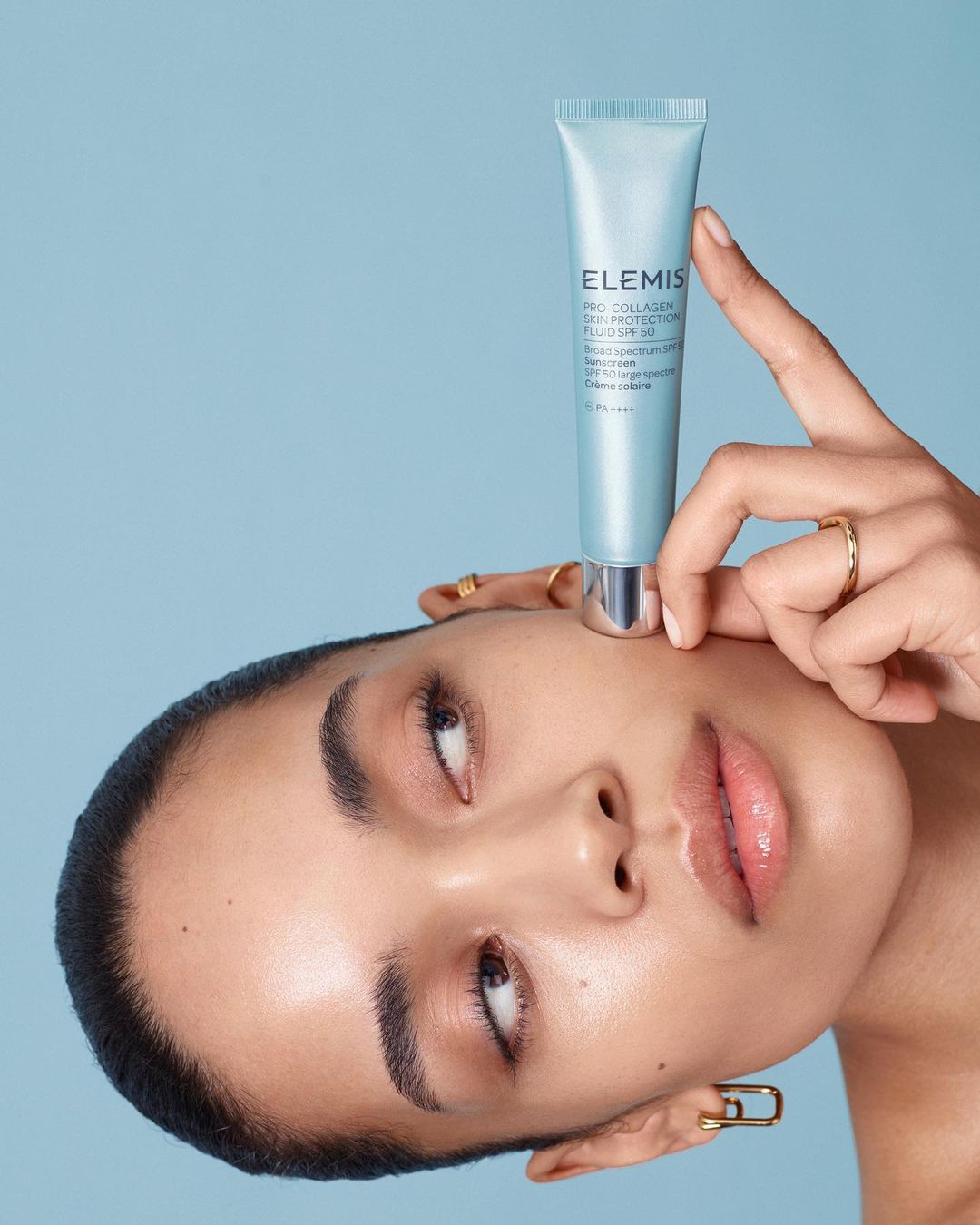 An adult woman with neutral facial expression holds a light blue Elemis skin protection fluid SPF 50 tube against a light blue background.