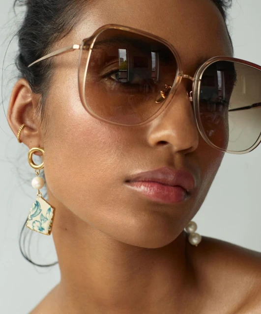 Close-up of an adult female model with tan skin wearing sunglasses and statement earrings in a neutral indoor setting.