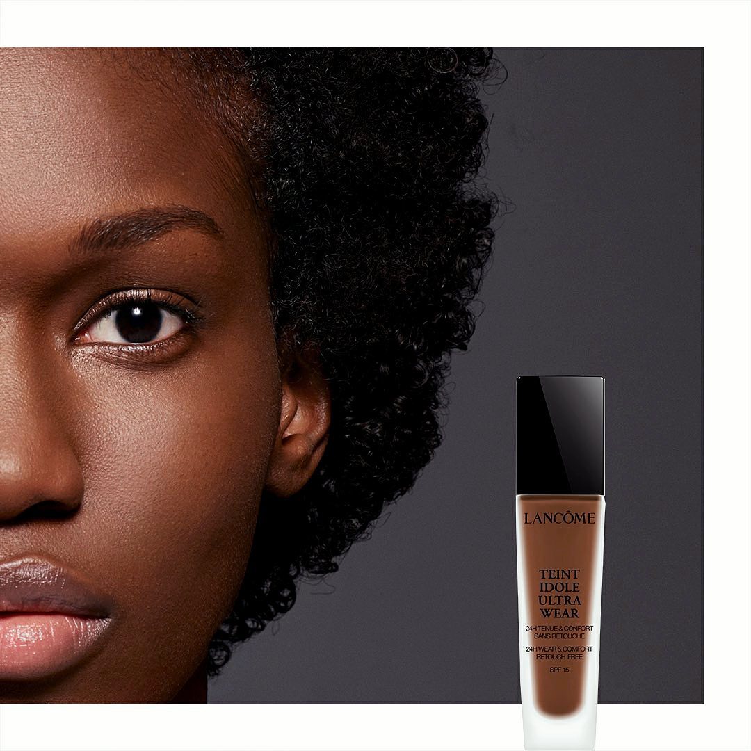 Close-up of a dark-skinned woman's face with a bottle of Lancome Teint Idole Ultra Wear foundation visible in the grey background.