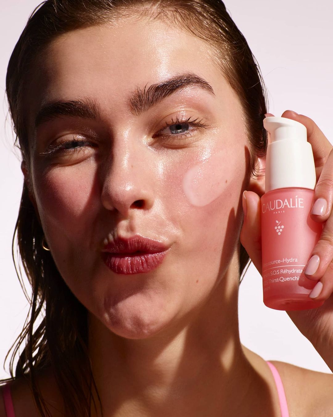 Close-up of a young woman with fair skin holding a pink Caudalie skincare bottle, showcasing dewy skin with light pink makeup.