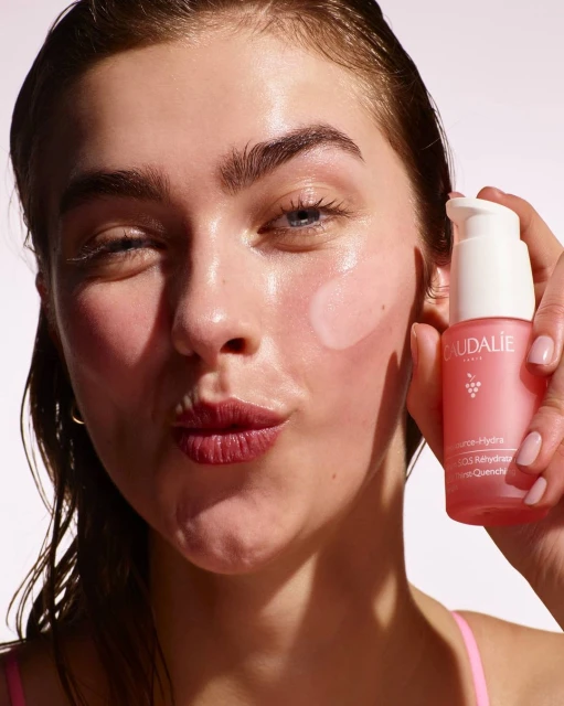 Close-up of a young woman with fair skin holding a pink Caudalie skincare bottle, showcasing dewy skin with light pink makeup.