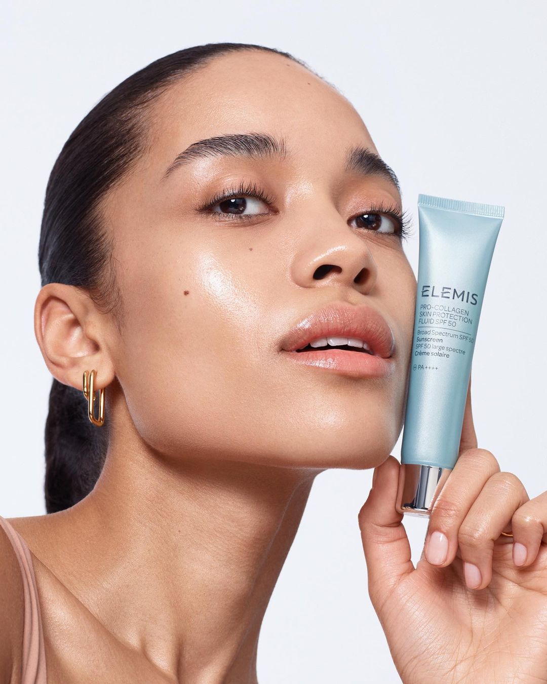Close-up beauty shot of a woman holding an Elemis skincare product tube in a brightly lit photo studio, showcasing skincare and beauty.