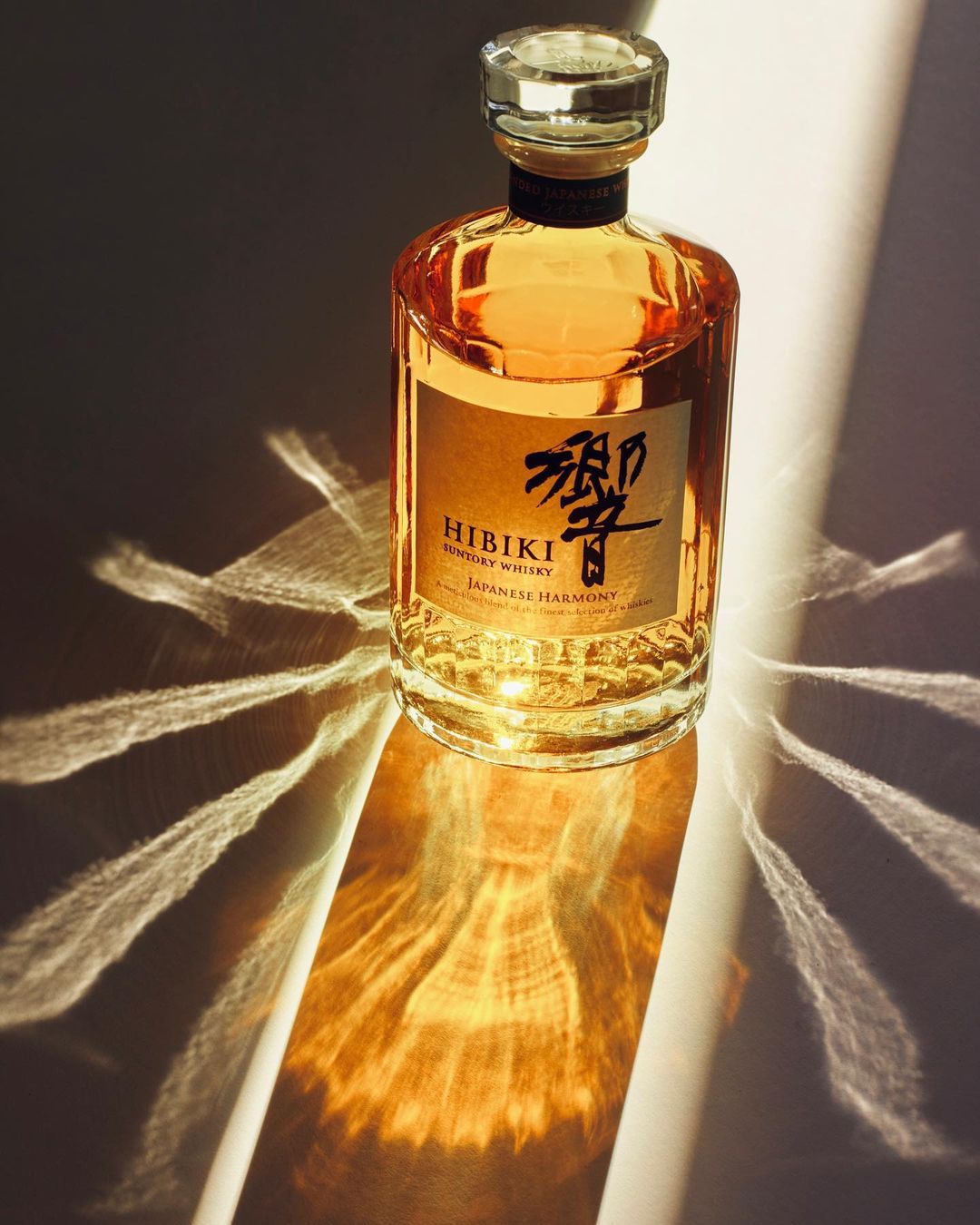 Bottle of Hibiki Japanese Harmony whisky on a reflective surface with refracted light patterns, showcasing its amber color and design.