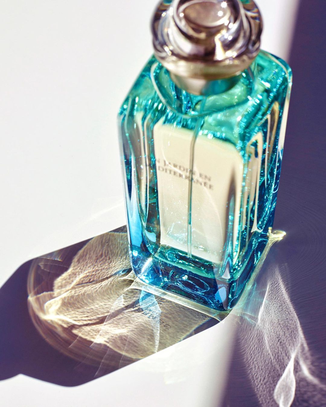 Turquoise glass perfume bottle with silver cap casting patterned shadows on white surface, product shot.
