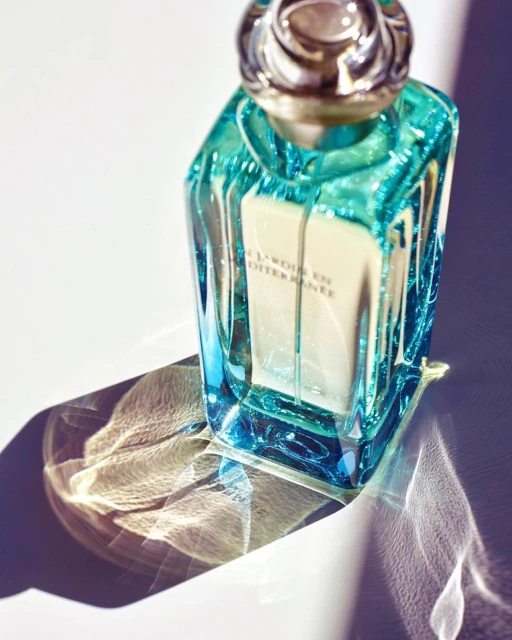 Turquoise glass perfume bottle with silver cap casting patterned shadows on white surface, product shot.