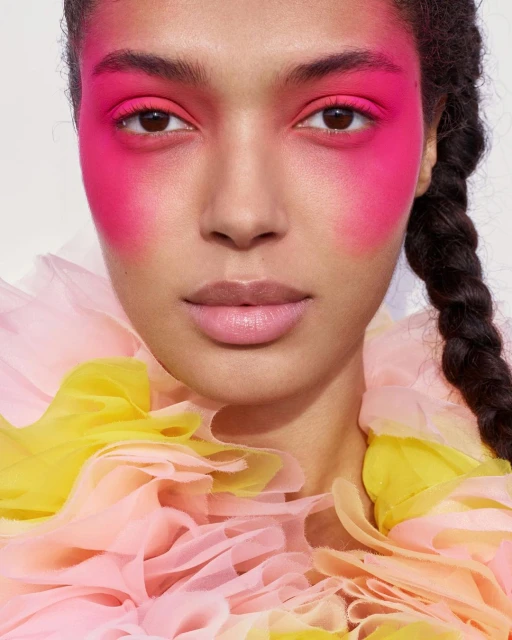 Close-up of a woman with pink eyeshadow and blush wearing a yellow and pink tulle ruffled top, posing against a white background.
