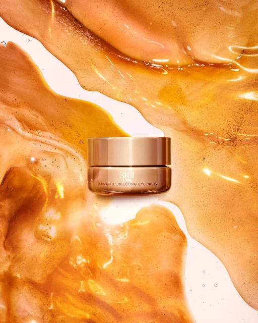 Eye cream jar by SK-II, in a gold colored container. The product is displayed on a background with streaks of gold liquid.