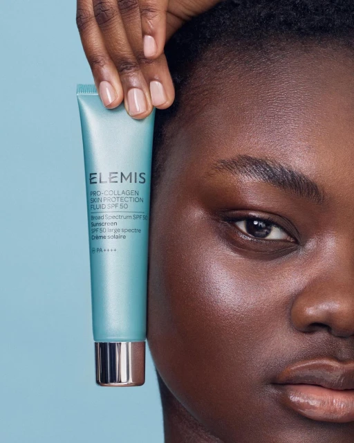 Close-up shot of a dark-skinned woman holding a tube of Elemis SPF 50 sunscreen, emphasizing skincare and sun protection against a soft blue backdrop.