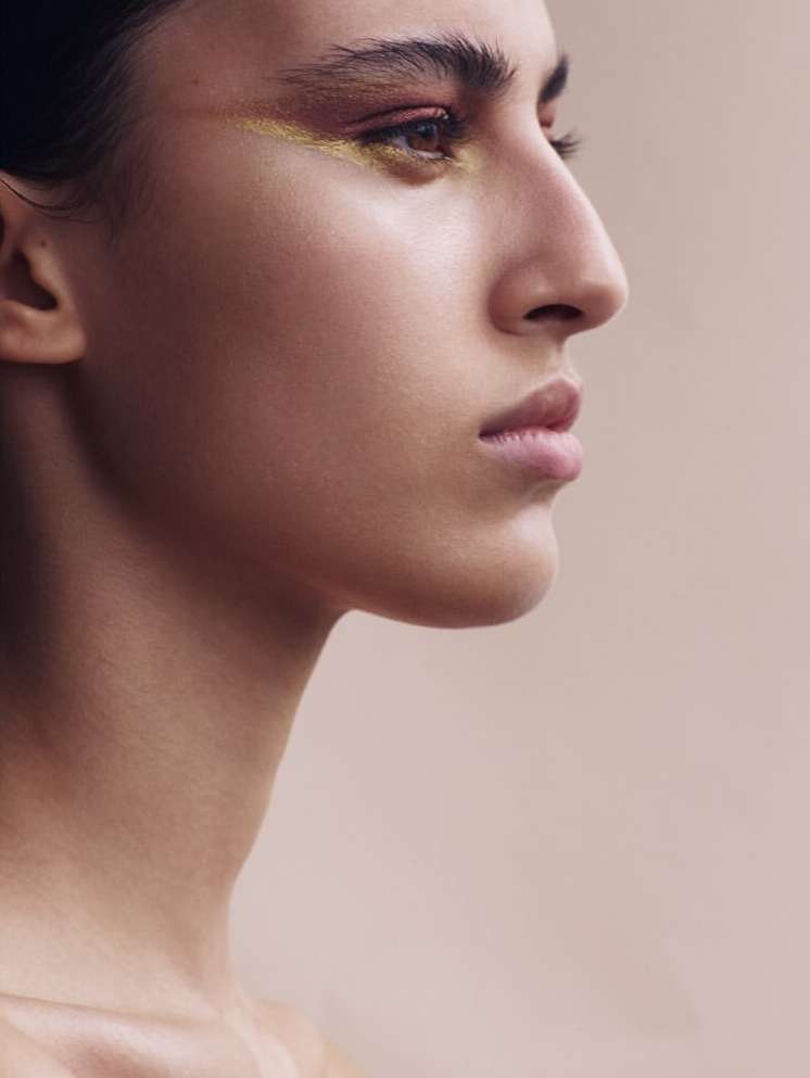 Profile of a woman with brown hair and gold eye makeup against a light neutral background, close-up shot of her face and shoulders.