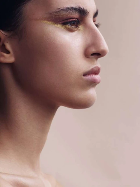 Profile of a woman with brown hair and gold eye makeup against a light neutral background, close-up shot of her face and shoulders.