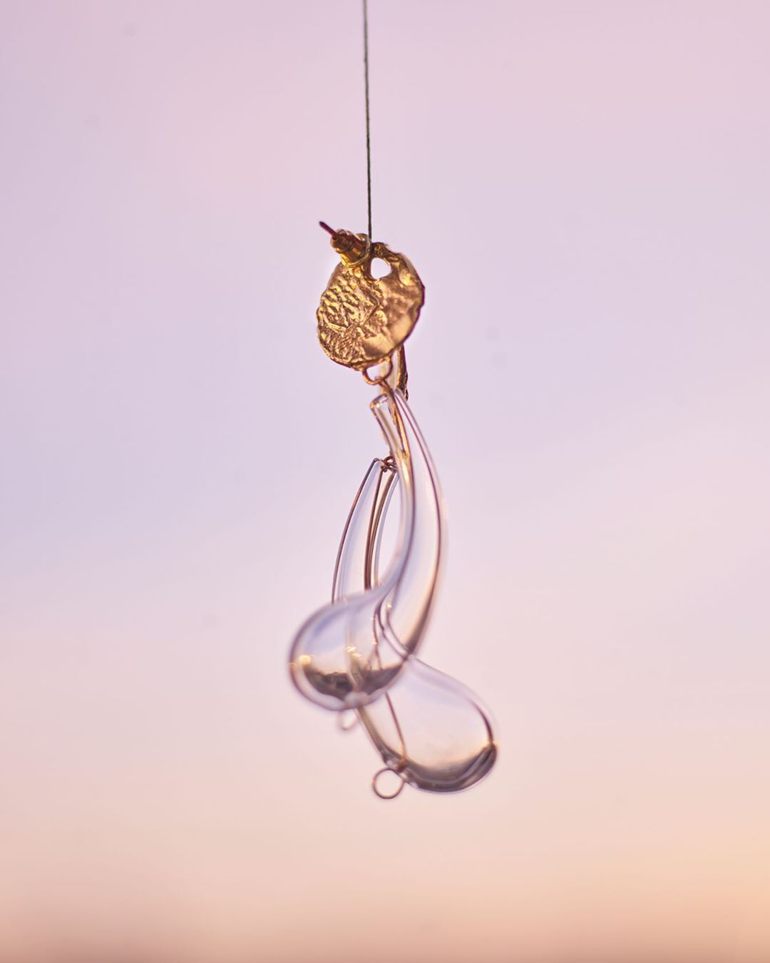 Macro shot of a handmade glass sculpture with golden elements hanging from a thin thread against a soft purple and orange gradient backdrop.