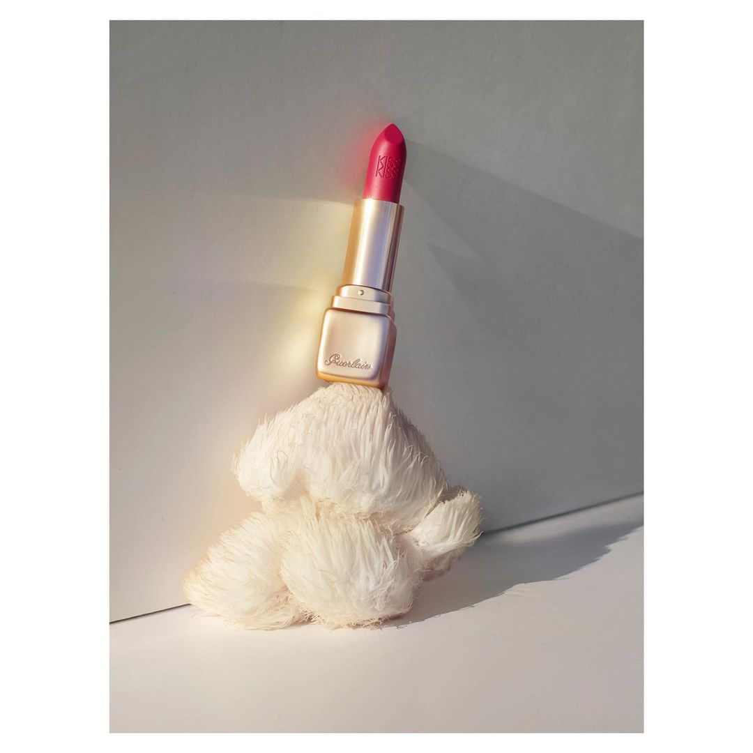 Guerlain lipstick tube in gold case with red lipstick sits atop a fluffy white prop against a minimalist white and gray studio backdrop.
