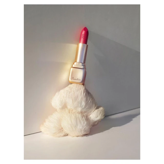 Guerlain lipstick tube in gold case with red lipstick sits atop a fluffy white prop against a minimalist white and gray studio backdrop.