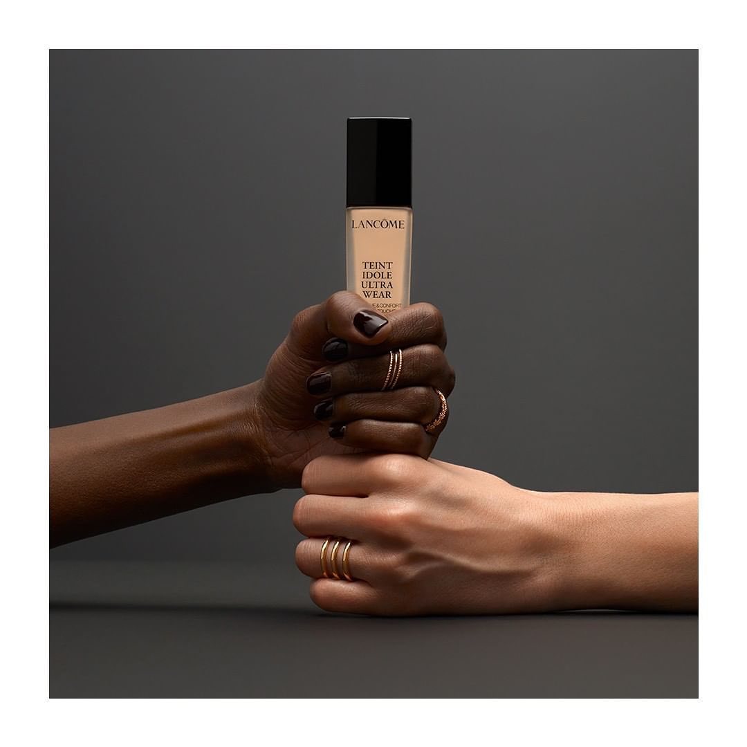 Lancôme Teint Idole Ultra Wear foundation bottle held between two hands, showcasing different skin tones in a beauty product advertisement.