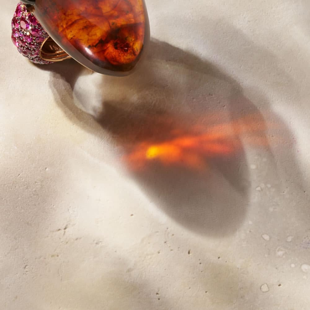 Amber gemstone ring with pink gems on a textured surface, casting a diffuse shadow with an amber-toned glow on a neutral background.
