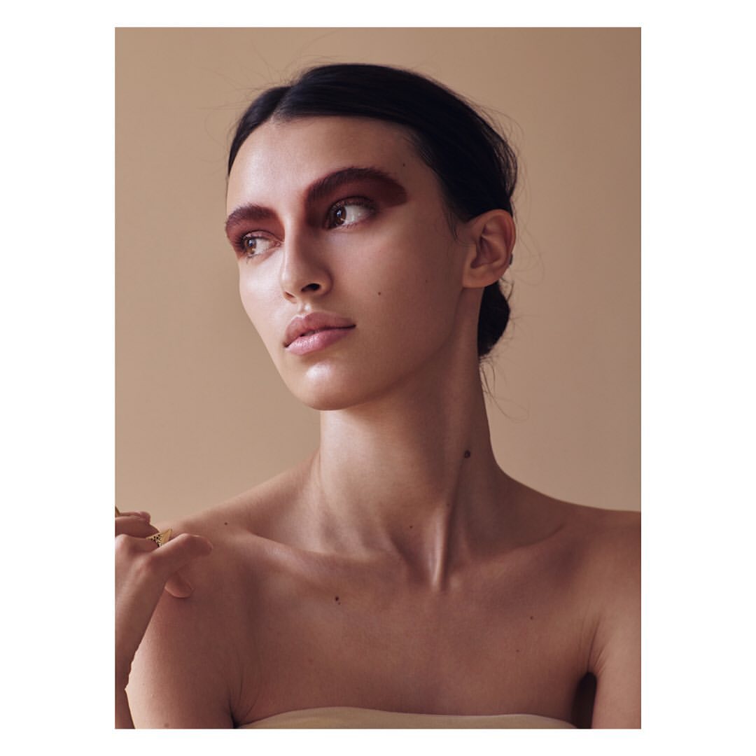 Close-up portrait of a young woman with brown eyeshadow, fair skin, and dark hair, wearing minimal clothing against a neutral backdrop.