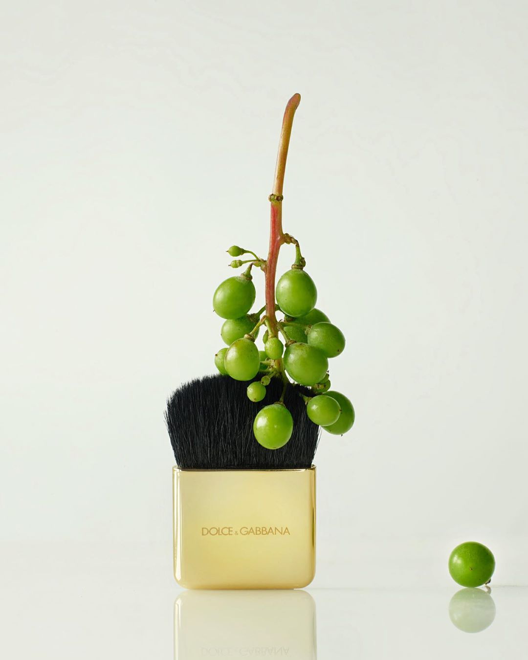 Dolce & Gabbana powder brush topped with a bunch of green grapes on a white reflective surface in a beauty product shot.