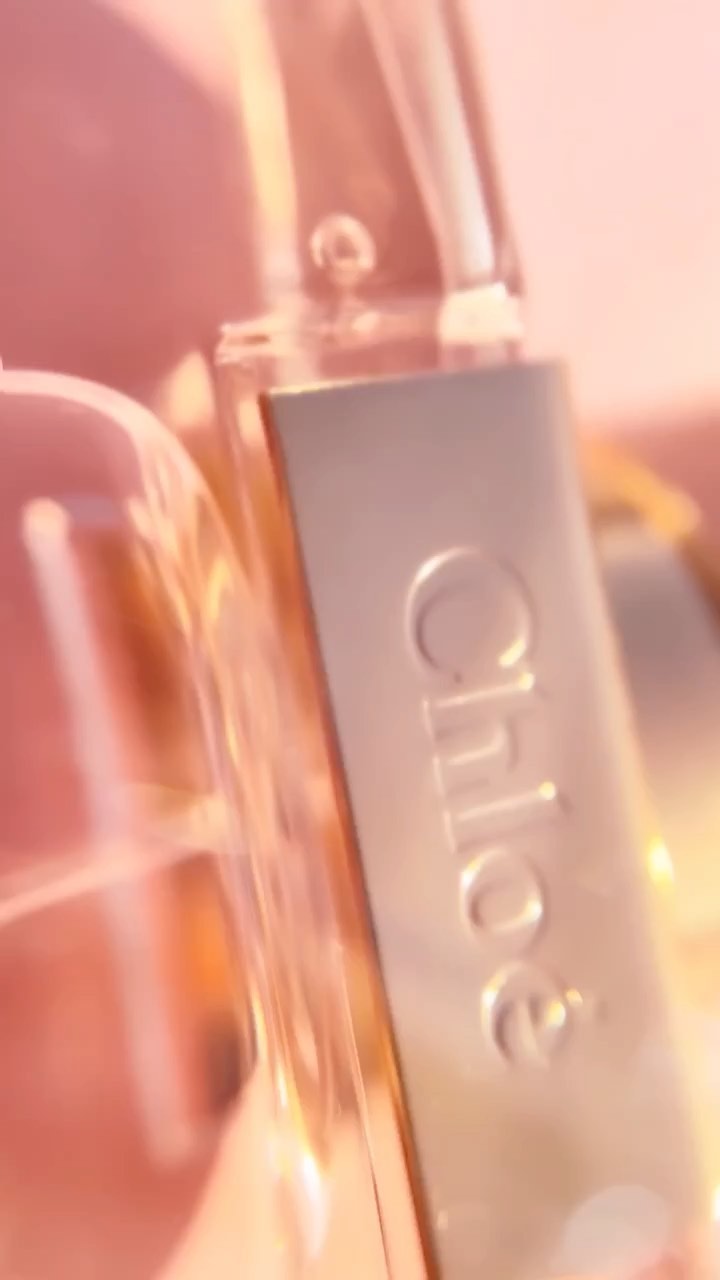 Close-up product shot of a bottle of Chloe perfume with a dropper, against a soft, diffused, pink-toned background and bright, diffused lighting.